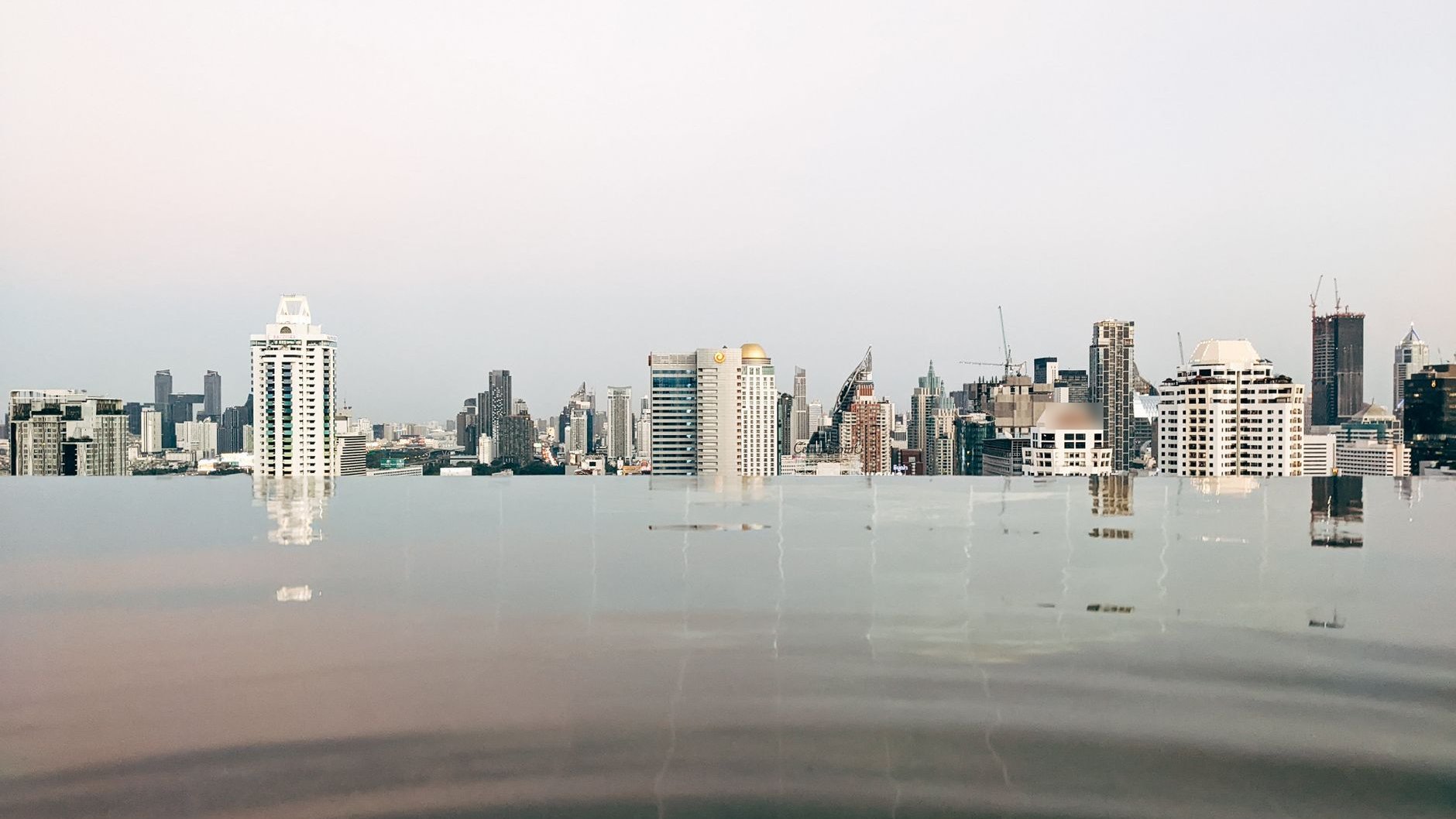 Bangkok high-rise skyline under a pale hazy sky, seen across the edge of a rooftop pool (illustrative stock photo)