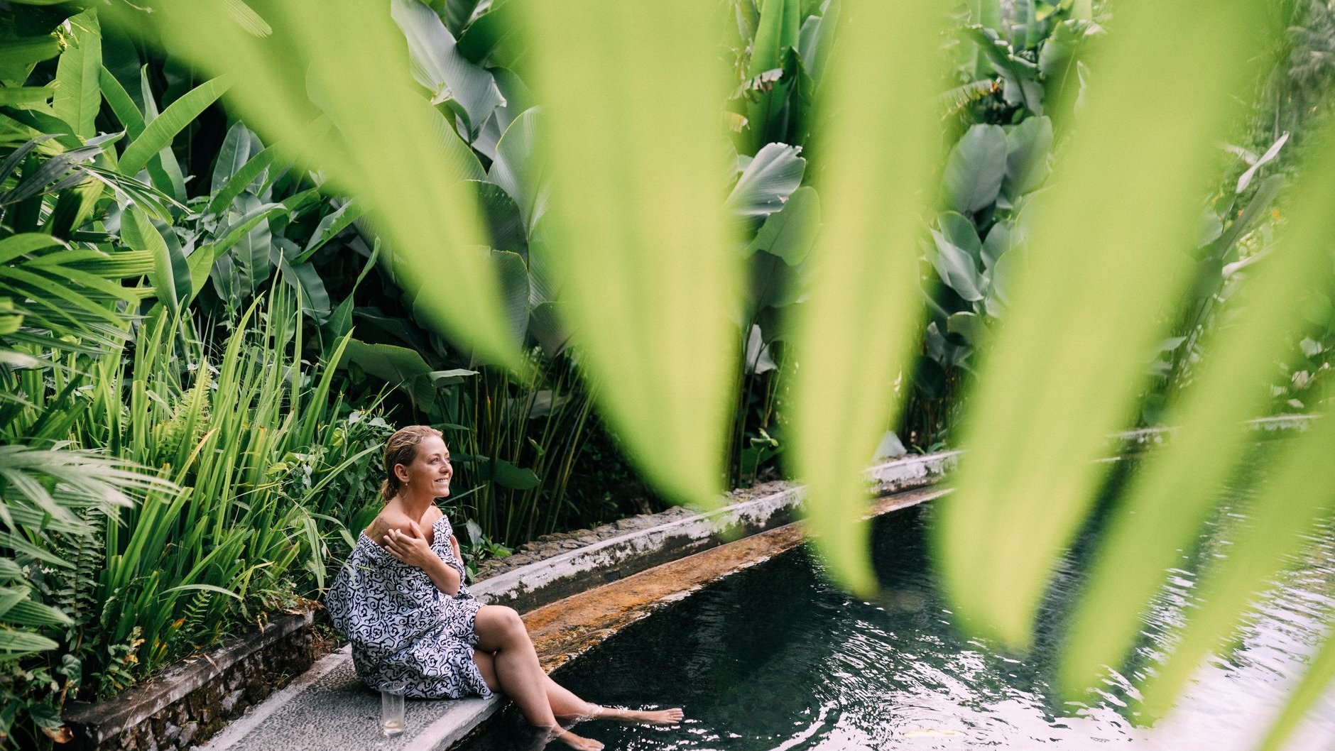 A woman wrapped in a sarong sits at the edge of a tropical pool with her feet in the water, surrounded by large green leaves (illustrative stock photo)